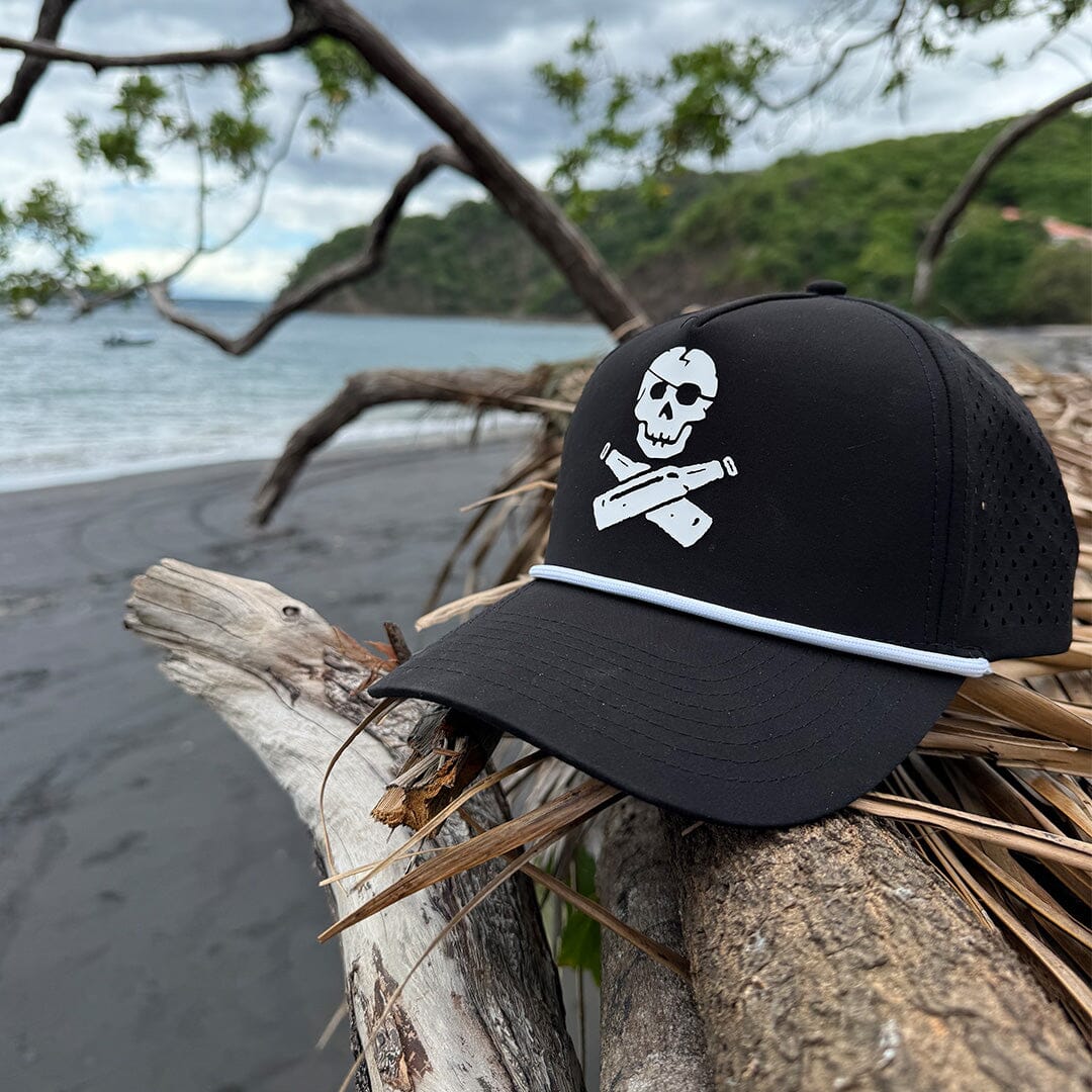 A black baseball cap with a white skull and crossbones graphic on the front, resting on driftwood on a beach with the ocean and green hills in the background.