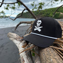 A black baseball cap with a white skull and crossbones graphic on the front, resting on driftwood on a beach with the ocean and green hills in the background.