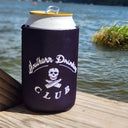 A Black neoprene can cooler sleeve with a white 'Southern Drinking Club' skull and crossbones logo, holding a white can with a gold lid, set on a wooden dock with a lake in the background.
